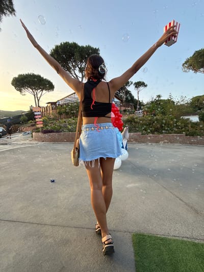 The image captures a rear view of a young woman with her arms outstretched, standing on a paved surface with scattered bubbles in the air. She is wearing a black crop top with orange straps, a light blue denim skirt with a frayed hem, and black sandals. A straw crossbody bag hangs from her left shoulder. In her right hand, she holds a red and white striped popcorn box. To her left, a stack of red, white, and blue balloons is visible, along with a white seal figure and more balloons. The background shows a landscape with trees, vegetation, and a building with signage. The setting appears to be outdoors, possibly a park or an event venue, with a clear sky overhead, suggesting it is daytime or early evening. The presence of balloons, bubbles, and a popcorn box indicates a festive or celebratory atmosphere. Based on the context provided, the location is the city of Sant Vicenç De Montalt, Spain.