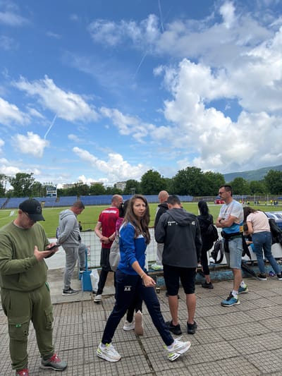 This image depicts an outdoor scene at a stadium in Sevlievo, Bulgaria, under a partly cloudy sky. In the foreground, a group of people are gathered, some standing and others walking. A man in a green tracksuit and black baseball cap is looking at his phone. Beside him, a man in a grey tracksuit stands with his hands behind his back. Further to the right, a woman in a blue and white jacket is walking, her face partially obscured. Other individuals are visible in the background, including a man in a red shirt and a man in a white t-shirt with a colorful design. The stadium seating, a sports field, trees, and a distant hillside are also visible. The ground is paved with square tiles.