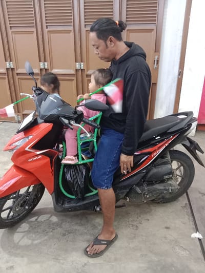 This image, taken in Kaliwungu, Indonesia, displays an adult male and two young children positioned with a red and black Honda Beat scooter. The adult male, wearing a black hooded sweatshirt, blue denim shorts, and dark grey flip-flops, stands to the left of the scooter, facing right. His dark hair is tied in a bun, and he holds a lit cigarette in his right hand, which rests on the scooter's black seat. A young child, dressed in a pink long-sleeved shirt, pink pants, and pink sandals, is seated in a green metal-framed child seat secured to the scooter's front footwell. This child holds a small red and white flag on a green stick. A second young child is partially visible behind the first child, also holding a similar red and white flag on a green stick. A black plastic bag hangs from the front of the green child seat. The scooter is parked on a light grey concrete surface. The background features a wall with two closed wooden louvered double doors. A vertical red and white stripe is visible on a wall section to the far right.