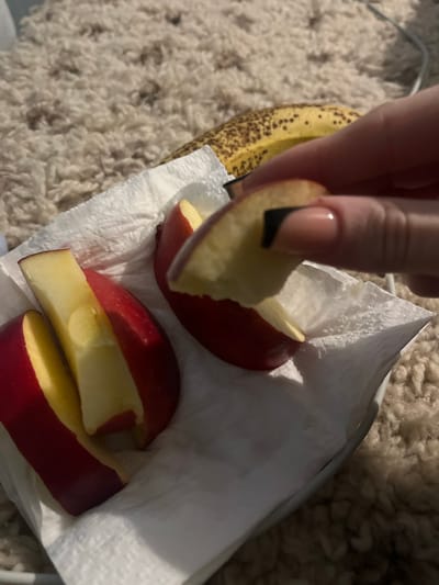 A hand with a beige and black fingernail holds a partially eaten slice of red apple. The apple slice has a bite taken out of it. Below, on a white paper towel, are two more apple slices that have also been partially eaten. A ripe banana sits next to the paper towel. The background is a fuzzy, light-colored rug. No indication of Blaskovo, Bulgaria is present.