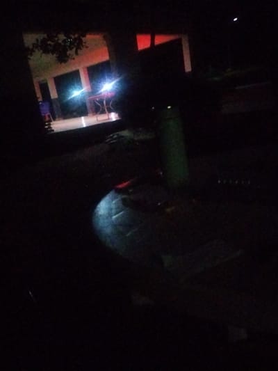 The image depicts an outdoor scene at night in Mangu Town, Nigeria. The setting appears to be a courtyard or paved area adjacent to a building. In the background, there's a building with illuminated windows showing a reddish-orange glow, possibly from interior lighting or decorative lights. Two bright white lights, potentially from car headlights or powerful lamps, are prominently visible, casting reflections and illuminating the scene.
In the foreground, a large, dark, circular object with a textured surface is visible, taking up a significant portion of the frame. Its exact nature is unclear due to the low light conditions and the image's resolution, but it could be a large pot, a decorative planter, or a piece of outdoor furniture. To the left of this object, a slender green cylindrical object, possibly a post or a narrow container, stands upright.
The overall mood is dark and somewhat mysterious, illuminated primarily by artificial lights. The time of day is clearly night, and there are no indications of the weather. The scene is likely a public or semi-public space, given the building and the paved area. No people are clearly discernible, and no text is visible in the image. The presence of the building suggests it might be a school, a community center, or a residential complex. The bright lights could indicate activity, or simply be ambient lighting for the area.