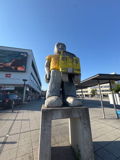 This image captures a statue of a person wearing a bright yellow vest and grey trousers, likely an outdoor sculpture. The statue is positioned in a public space, possibly a city square or sidewalk, with a modern building featuring large windows and signage in the background. A bus stop shelter is also visible. The ground is paved with large square tiles, and shadows suggest it is daytime.

The text visible on the building includes "BOARD27", a phone number "07142/77182-0", and a business name "POINT OF CUT Bietigheim". Another sign reads "Itur". These details suggest the location is Bietigheim-Bissingen, Germany.

The scene appears to be a typical urban environment on a clear, sunny day. There are a few people visible in the distance, some walking and one sitting. The overall atmosphere seems calm and ordinary, with the prominent statue serving as a point of interest in the urban landscape. The statue itself appears to be made of a rough, textured material, giving it a somewhat raw and artistic quality. The photograph is taken from a low angle, emphasizing the scale of the statue and the surrounding architecture.