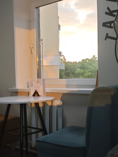 The image depicts the interior of a cafe in Svietlahorsk, Belarus, looking out of a window. A small, round white table with a marble-like pattern sits in the foreground, supported by black legs. On the table, a menu or brochure is propped up, displaying text and a graphic of a mountain. A radiator is visible beneath the windowsill. To the right of the table, a teal upholstered chair is partially visible.

Through the window, a building with a cream-colored facade and visible air conditioning units is seen, along with a stand of green trees and a cloudy sky. A white window frame encloses the view. A part of a sign with the text "TAK" is visible on the right edge of the image. The lighting suggests it is either early morning or late afternoon, with warm, golden sunlight casting shadows. No people are present in the image.