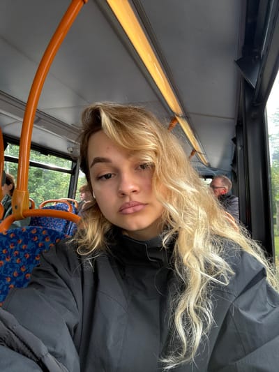 The image shows a young woman with long, blonde, wavy hair wearing a black jacket while riding a bus in Crail, United Kingdom. She looks directly at the camera with a neutral expression. Her hair covers most of the right side of her face. The bus's orange handrail is in the foreground. Another person sits in the background, partially visible on the right side of the frame. The bus appears to be in motion, and the woman appears to be taking a selfie. A window to the left of the woman shows green trees and foliage, and a window to the right shows greenery and a house in the distance.