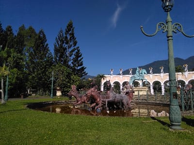 The image depicts a park scene in Batulawang, Indonesia, under a clear blue sky with a single wispy cloud.
In the foreground, manicured green grass covers the ground, leading to an irregularly shaped pond with dark, still water. Situated within this pond is a multi-figure sculpture group, cast in a reddish-brown material, depicting a quadriga (four horses) pulling a chariot. Two human-like figures are discernible within the chariot assembly. A tall, ornate, dark green lamppost with two curved arms stands prominently on the far right, partially cropped. Another similar lamppost is positioned further left within the grassy area.
The midground features a large, light-colored building with a classical architectural style. Its facade is characterized by a series of arches supported by columns or pilasters, spanning the length of the visible structure. The building's parapet is adorned with uniformly spaced decorative elements, likely urns or small statues, appearing light-colored with gold accents. Centrally positioned in front of this building, atop a pedestal, is a large equestrian statue depicting a figure on a rearing horse, cast in a material with a distinct patinated blue-green hue. Dense green foliage, including tall coniferous trees and smaller bushes, flanks the left and center-left portions of the midground.
The background reveals a distant, dark green mountain range visible beneath the clear blue sky, extending across the horizon. The entire scene is brightly illuminated by daylight.