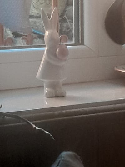 A white ceramic figurine of a bunny holding a pink egg stands on a white ledge, possibly a windowsill. The bunny is wearing a dress and has long ears. In the background, a window is visible with what appears to be a blurred outdoor scene. The image is a close-up shot, with the focus on the figurine. The setting is likely indoors, given the presence of the ledge and the way the light is diffused. The context of Kostenets, Bulgaria, is not visually apparent from the image itself and would require external information.