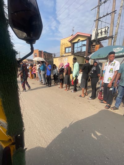 In Akowonjo, Nigeria, on a paved street, a diverse group of individuals stands in a line. The scene is captured from a low angle, with a portion of a yellow vehicle's exterior, covered in green artificial turf, occupying the left foreground. Behind the vehicle, a man in a vibrantly patterned outfit walks away from the camera.

The main focus is on the line of people stretching into the distance. The individuals are of varying ages and genders, dressed in casual attire. Some are looking forward, others are conversing with those next to them. One woman in the middle of the line holds a white umbrella. Behind the line of people, buildings with colorful facades and exposed electrical infrastructure are visible. The sky above is partly cloudy. A green umbrella with the text "MORNING FRESH" and "CHEN CARE EX" is situated to the right of the line. The overall impression is of a busy street scene in a Nigerian city.