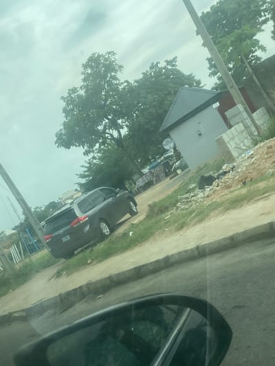 This media file captures an outdoor street scene, viewed from inside a vehicle, with the side-view mirror partially visible on the right. The image, originally rotated, shows a paved road extending towards the left and a parked dark-colored car on its side. The car, possibly a sedan or small SUV, has a partially visible rear license plate that appears to read "000 87Q."

Beyond the road, there is a grassy verge and a row of buildings interspersed with abundant green trees. One prominent building in the mid-ground features a dark, corrugated metallic roof and light-colored walls. Near these structures, a circular sign is visible, clearly displaying the word "EATS," with further, unidentifiable text beneath it. A tall utility pole or signpost stands on the far left of the scene. The sky is uniformly overcast, indicating a cloudy daytime setting. No individuals are visible in the image. The environment suggests a typical street in a populated area, aligning with the provided location context of Lugbe, Nigeria.