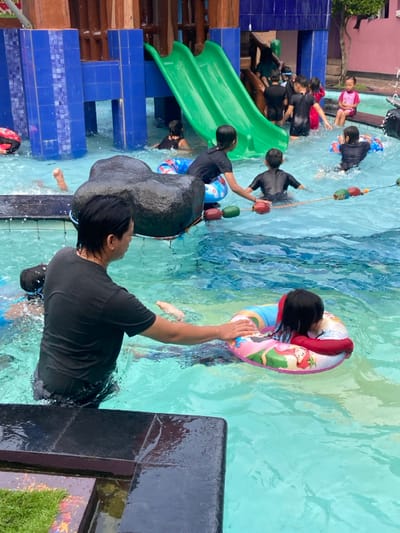 A man in a black shirt is in a swimming pool, helping a young girl with a pink and white float. Several other people are in the pool. There are blue tiles around the edge of the pool and a large rock in the middle of the pool. There are green water slides in the background. This scene appears to be a public swimming pool in the city of Kecamatan Tanggulangin, Indonesia, which is located at coordinates (latitude: -7.497, longitude: 112.684).