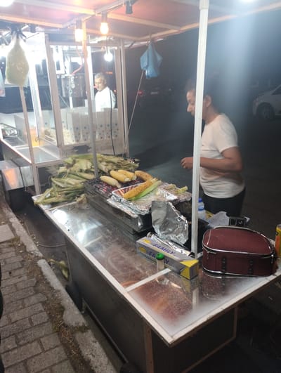 A man is seen working at a food cart selling corn. The cart is illuminated and appears to be outdoors at night. The man, wearing a white t-shirt, is bent over the cart, presumably preparing or serving food. The cart is laden with corn on the cob, some of which are on a grill, and other food items are visible inside the cart. There are also bags of popcorn and what appears to be cotton candy. A maroon bag is placed on top of the cart. The scene is set on a brick-paved area, with a street visible in the background. The overall impression is that of a street vendor operating in a Greek location, likely Neoi Epivates. There is some text visible on a yellow box which reads "FAMILY".