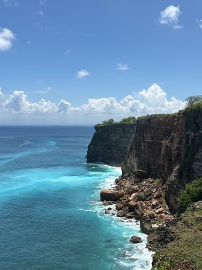 The image depicts a view from above of Uluwatu, Indonesia. A rugged cliff face, topped with green vegetation, plunges down to the turquoise ocean. Waves crash against the rocks at the base of the cliff. The sky is blue with scattered white clouds.