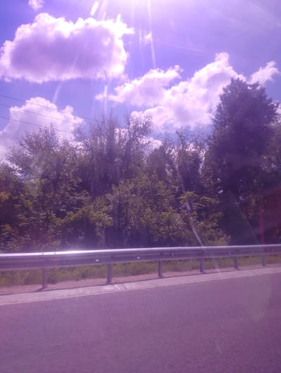 The image depicts a roadside scene with a metal guardrail separating the road from a treeline. The road surface is a muted purple, suggesting it's either asphalt with a color filter or has been affected by light conditions. Beyond the guardrail, a dense collection of green trees forms a natural barrier, their leaves lush and full. In the background, the sky is a vibrant purple-blue, filled with fluffy white clouds. Sunlight streams down from the upper right corner of the image, creating a lens flare effect that adds a bright, ethereal quality to the scene. The sun's rays are visible as streaks of light, illuminating the clouds and sky. A few power lines are visible against the sky, stretching horizontally across the upper portion of the image. The overall impression is of a sunny day, possibly late afternoon given the warm light, with a clear view of the natural landscape next to a road. There are no people or specific objects other than the guardrail and trees. The location is identified as Otaci, Moldova.