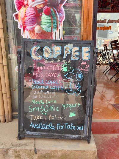 A black, double-sided, A-frame chalkboard menu board stands on a light brown concrete step outside a retail establishment in Sa Pa, Vietnam. The board displays hand-written menu items in various chalk colors, including "COFFEE" in large decorated letters, followed by "Eggs coffee," "Milk coffee," "Black coffee," "Coconut coffee," "Faint coffee," "Hot Chocolate," "Matcha Latte," "Smoothie," "Juice," "Hot tea," and "yogurt." The bottom of the sign states, "Available For Take out." Decorative chalk drawings include a light blue Snoopy-like dog character, a green frog, a pink hot beverage cup, a green iced beverage cup, and small yellow stars.
To the immediate left of the chalkboard is a glass storefront window featuring a large, colorful decal of an ice cream cone with three scoops (pink, green, and white soft-serve), topped with raspberries and chocolate drizzle. Reflections on this window include text such as "GSCL 14-04/2025" and "KEM 40 X 1.0". Several vertical bamboo poles are visible along the far left edge. To the right, a dark wooden door frame separates the left window from another storefront window. This right window reflects an outdoor seating area with dark, foldable tables and chairs. Reversed text, including "Souvenirs, Handmade," is visible in the reflection. The ground immediately beyond this doorway is tiled with brown and tan geometric patterns. A red-painted concrete step extends into the right foreground. A black cable, resembling a USB cable, dangles from beneath the menu board onto the concrete step. No individuals are present in the image.
