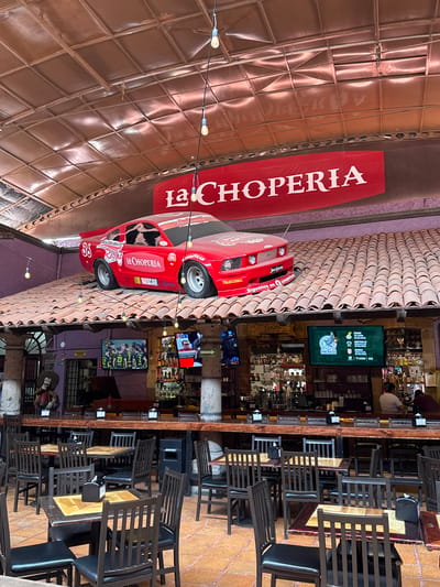 The image depicts the interior of "La Choperia," a bar in San Miguel de Allende, Mexico. A red race car replica hangs from the ceiling alongside a banner with the bar's name. Strings of lights extend across the ceiling. The bar area features multiple shelves of bottles and several television screens displaying sports broadcasts. Seating consists of wooden tables and chairs. A wooden structure and terracotta tiles are part of the architectural elements.
