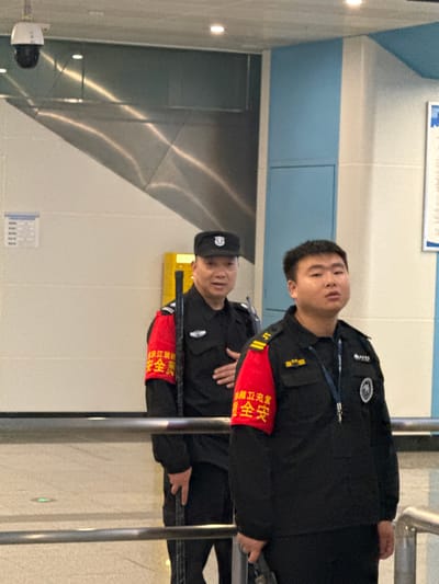 This media file, captured in an indoor setting likely in Shuangfu, China, depicts two men, presumably security personnel, standing guard. The image appears to be rotated 90 degrees clockwise from its natural upright orientation.

The primary subject is a young man in the foreground, looking directly at the camera. He wears a black uniform jacket with a badge on his right lapel and a red armband on his left arm. Behind him, another man, older and wearing a black cap, is partially visible, also in a black uniform and a red armband. Both men's armbands bear Chinese characters. They are positioned behind a series of vertical metal bars, suggesting a controlled access point or barrier.

The setting features a bright, clean environment. The background consists of a wall with a light blue stripe running horizontally across its lower section, topped by a white wall. To the left, there's a metallic, reflective wall with subtle light reflections. A small, dark security camera or device is mounted on the lower left wall. Another small, unreadable white sign is visible on the lower left wall, and a larger white sign with blue elements is partially visible on the upper wall.

The visible text on the red armbands is "治安巡逻安全" (Zhì'ān Xúnluó Ānquán), which translates to "Public Security Patrol Safety" or "Security Patrol Safety." The uniforms, armbands, and the presence of barriers indicate an official security or public service role within a public building such as a transportation hub or facility entrance. Both individuals maintain a neutral expression, with the man in the foreground appearing observant. No specific action beyond standing guard is taking place. The indoor lighting suggests no particular time of day.