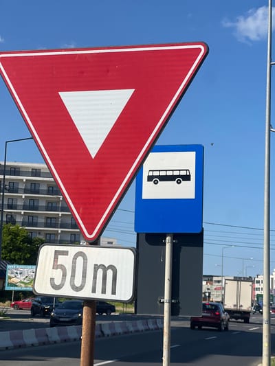 The image shows a street scene in Năvodari, Romania, on a clear, sunny day. In the foreground, three traffic signs are visible. The topmost sign is a red and white yield sign, shaped like an inverted triangle with a white triangle inside. To its right, a blue sign with a white silhouette of a bus indicates a bus stop. Below the yield sign, a rectangular white sign displays "50m" in black numbers and letters, likely indicating a distance. The signs are mounted on metal poles, one of which appears weathered and rusty.

In the background, a multi-story building with numerous windows and balconies is visible, suggesting a residential or commercial area. To the left, a billboard advertises "MEGA KILIAN RESORT" with an illustration of modern apartments and trees. The street is paved and marked with white lines. Several cars are on the road: a red sedan, a dark SUV, and a truck with a white cargo box. Traffic lights and street lamps are also present. The sky is a vibrant blue with a few wispy white clouds and a small bird in flight. The scene conveys a typical urban or suburban environment with active traffic.