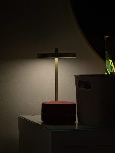 A table lamp with a thin, brass-colored stand and a rectangular light fixture sits on a dark red, circular base. The lamp is positioned on a modern, white dresser with a glass top, reflecting a faint green hue. To the right of the lamp, a light-colored storage basket with cut-out handles is visible. The background consists of a textured, off-white wall, illuminated by the lamp's glow, creating a soft halo effect. The overall scene is dimly lit, with shadows dominating the edges of the frame. The image was taken in Oeiras, Portugal.