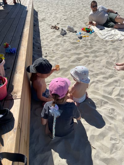 In Zelenogradsk, Russia, on a sunny day at the beach, three young children are playing in the sand near a wooden deck. Two children are seated on the sand, facing away from the viewer. The child on the left is wearing a black baseball cap and is holding a small cup. The child on the right is wearing a pink hat. A third child, an infant, is sitting between them, facing the same direction, wearing a white sun hat with a cherry pattern and a diaper. In the background, a man is lying on a beach towel. Beach toys, sandals, and a bucket are scattered on the sand. The environment is a sandy beach with a wooden deck.