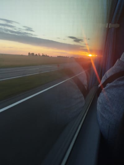 The image captures a sunset viewed from the window of a vehicle, likely a bus, traveling on a road. The sun is setting on the horizon, casting an orange glow across the sky and reflecting off the window. In the distance, a flat landscape with trees is visible. The interior of the vehicle is partially visible, showing a person wearing a grey hooded garment and a seatbelt. The road has white lane markings. The overall impression is that of a journey at dusk through a rural area.