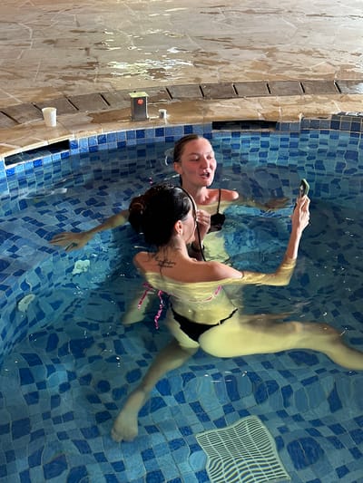 Two women are captured in a vertical, overhead shot while relaxing in a tiled pool. The pool's interior features a checkerboard pattern of blue and lighter blue mosaic tiles.
One woman, positioned further away from the camera, has lighter hair and is floating on her back with her head partially out of the water. Her eyes appear closed or squinting, and her mouth is slightly open, conveying a sense of relaxation. She is wearing a black bikini top.
The second woman, closer to the camera and mostly submerged, has darker hair tied back. She is holding a smartphone with a prominent green case above the water with her right hand, seemingly taking a photo or video. A script tattoo is visible on her left shoulder/upper back. She appears to be wearing a light-colored (possibly white or pink) bikini top with pink straps. Both women's submerged limbs are visible through the clear water, which shows ripples and reflections.
The pool's edge, visible along the left side of the frame, is constructed from wet, beige and brown mosaic tiles or stone. Mounted on this wall are a small metallic fixture with a red light and a white cup. A white, grid-patterned grate, likely a drain or overflow, is visible on the pool floor towards the right.
The scene depicts a leisure activity in what appears to be an indoor or covered pool setting, possibly a resort or spa in Mu Cang Chai, Vietnam. The overall impression is one of enjoyment and relaxation.