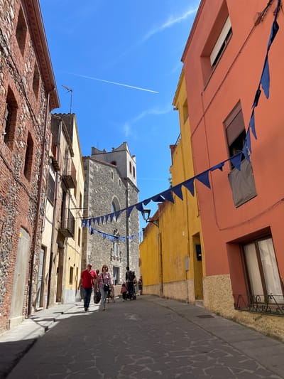 A low-angle shot in Hostalric, Spain depicts a narrow street scene with buildings of varying colors lining the sides. The buildings are constructed with brick and stone. Blue triangular banners are strung across the street overhead. Several people are walking along the street, including a man in a red shirt. A stone church tower is visible at the end of the street. The sky is blue, with a jet contrail visible. The street is paved with cobblestones.