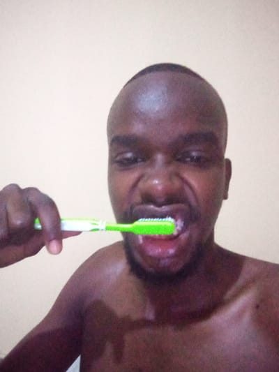 A dark-skinned, shirtless man is vigorously brushing his teeth with a green toothbrush. His face is close to the camera, showing his mouth open with toothpaste and water. The man's expression is intense, with furrowed brows and a slightly contorted face. The background is a plain, light-colored wall. The lighting is bright, highlighting the toothbrush and the man's wet skin.