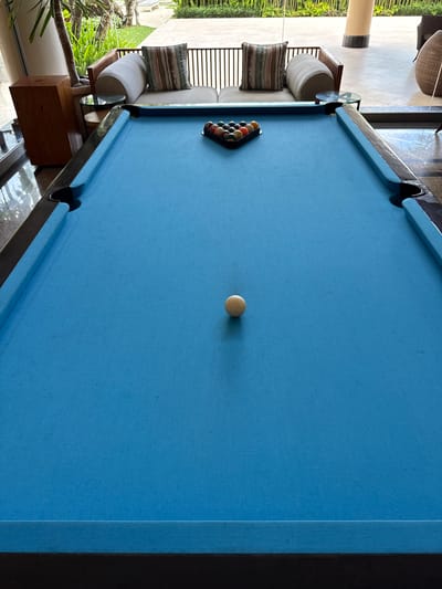 This image captures a pool table with a light blue felt surface. The billiard balls are arranged in a rack near the left edge, while the cue ball sits in the center of the table. The table's trim is dark, and its pockets are visible at various points along the edges. Adjacent to the table, a cushioned seating area with pillows is present. Beyond that is an outdoor area with landscaping and a glimpse of architecture. Based on the surroundings and the context provided, the location is Nusa Dua, Indonesia.