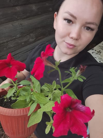 In this close-up, medium shot, a young woman is holding a potted plant with bright red petunias. She is positioned in the lower right quadrant of the image, with her face angled towards the camera and a slight smile. She has dark hair pulled back, and is wearing a black t-shirt. Her makeup includes dark eyeliner and false eyelashes. The petunia plant is in a reddish-brown plastic pot filled with dark soil. Several vibrant red flowers are in bloom, along with green leaves and stems. One unopened bud is also visible.
The background consists of weathered, dark wooden planks, suggesting an outdoor setting, possibly a rustic porch or fence. The lighting appears natural, indicating daytime. The overall mood is pleasant and casual, hinting at a moment of personal enjoyment in nature. No visible text is present in the image. The location is identified as Kuyeda, Russia.