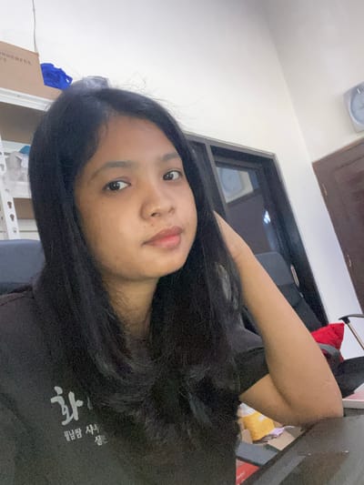 A young woman with long, black hair is the primary subject. She is fair-skinned and has dark eyes. She is wearing a dark t-shirt. Behind her is a partially visible interior space, including a closed dark wood door, a light-colored wall with a clock, and partially visible shelves or furniture with objects on them. Negara, Indonesia is not visibly present in the image, so location accuracy is not determinable.