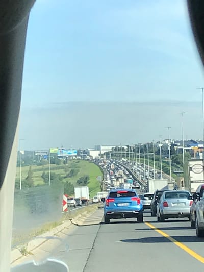 Heavy traffic stretches into the distance on a highway, with many cars packed closely together. A grassy area and billboards flank the side of the road. A blue car is visible in the foreground, along with other vehicles. Overhead streetlights are present. The sky is light blue, suggesting it is daytime.