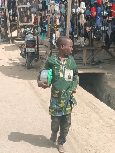 In Hotoro South, Nigeria, a young male child is depicted walking barefoot on an unpaved, light brown dirt path during daylight hours. He wears a knee-length green tunic adorned with yellow and blue geometric patterns, including a light-colored triangular pocket on the right chest, over dark trousers. In his left hand, he carries a cylindrical container, possibly plastic, with a bright green lid. He is looking towards his left, with his body angled slightly right. In the background, to the left of the child, a black motorcycle with license plate "KMC 67YH" is parked near a wooden stall. This stall, and another partially visible on the right, display numerous pairs of sandals and slippers in various colors (black, white, red, blue, green, brown, tan), some bearing brand-like emblems, hanging from wooden frames. A concrete or dirt drainage channel runs parallel to the child's path on the right side of the frame. Strong shadows indicate direct sunlight.