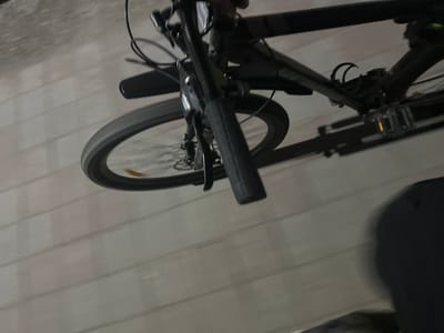 This is a close-up, low-angle shot of a bicycle, with the camera angled upwards. The bicycle is predominantly black with some silver accents. The handlebars and front wheel are clearly visible, with a hand grip on the right side. The bicycle has a fender over the front wheel. A portion of the bicycle's frame and pedals are also in view. The background appears to be a paved surface with horizontal lines, possibly a sidewalk or road. The lighting suggests it is either evening or nighttime, or an indoor setting with artificial light. There is no visible text on the bicycle or in the background. The scene is static, with no specific activity or event taking place other than the bicycle being stationary.