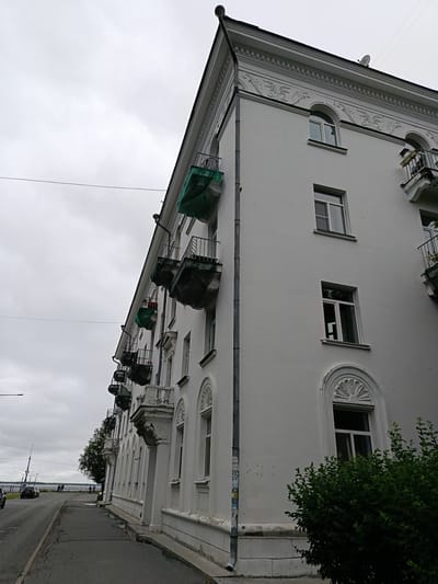 The image shows a white, multi-story apartment building with a distinctly European architectural style, featuring ornate detailing along the roofline and around the arched windows. Several balconies jut out from the building, some covered with green tarps, suggesting potential ongoing construction or maintenance. The building faces a street with a sidewalk and a body of water in the distance, likely a river or bay, given the location context of Arkhangelsk, Russia. The sky is overcast and gray, indicating a cloudy day, possibly with light rain or fog. A few cars are visible on the road, and a few people are seen walking along the waterfront promenade. The overall mood is somewhat somber due to the weather, but the grandeur of the building hints at a historical and established urban setting. No discernible text is visible in the image.