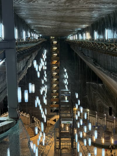 The image depicts the interior of the Turda Salt Mine in Boj-Cătun, Romania. The mine's rough-hewn walls are visible, illuminated by rows of fluorescent lights along the upper reaches. Suspended below are platforms and structures adorned with numerous horizontally-oriented light fixtures resembling glowing oblongs. Some platforms appear to have metal scaffolding. A green mesh netting is visible in the foreground. The mine's interior appears vast and multi-tiered.