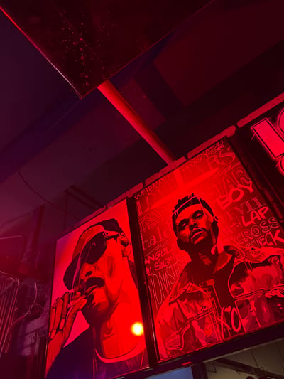 The image shows three illuminated signs under what appears to be a ceiling, with a red hue dominating the scene. The signs feature artwork including portraits and hand-drawn-style text. The text includes phrases such as "WE DUIN", "TRADE FOR", "ANGELS ALL SINGIN'IN", "STAR", "DEVIL ON MY LAP", and "ACROSS ON MY NECK". There is a dark, reflective surface on the left side of the image, reflecting the light. The location is possibly a bar or club environment, and is not identifiable as Linghe, China.