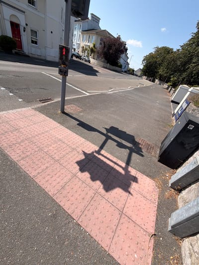 This image depicts a street scene in Torquay, United Kingdom, with a focus on a pedestrian crossing. A traffic light pole stands prominently, displaying a red pedestrian signal. The crossing itself is marked with tactile paving, characterized by rows of raised circular studs on pink tiles. The surrounding environment includes asphalt roads, a storm drain, and a sidewalk. In the background, residential buildings with white facades and red-tiled roofs are visible, along with mature trees and a clear blue sky. The sun casts long shadows, indicating a sunny day. The overall impression is that of a quiet urban street.