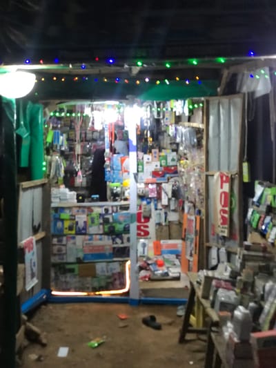 The image shows the front of a small shop in Galadimawa, Nigeria. The shop is packed with various items, seemingly electronics and accessories, densely arranged on shelves and walls. A string of colorful lights is strung along the shop's entrance. The ground outside is dirt. A blue metal frame partially outlines the right side of the shop's entrance. A single shoe lies on the ground near a table outside the shop.