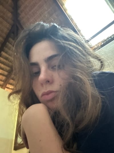 A low-angle selfie captures a person with long, tousled brown hair, resting their head on their arm. Their face is tilted, revealing slightly closed eyes and a neutral expression. Light enters from a window above, illuminating the hair and upper portion of the face. A ceiling constructed of natural materials is visible in the background, adding texture and depth. The overall composition creates a relaxed and intimate feel.