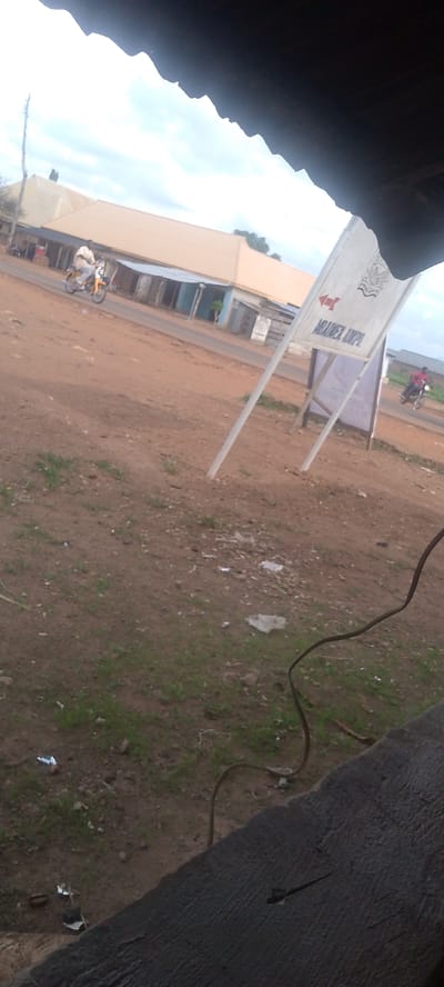 A man rides a yellow motorcycle on a dusty road in Adikpo, Nigeria. He is wearing a light-colored shirt and light-colored pants. The road is lined with low buildings that have corrugated metal roofs. A white signpost with a red arrow pointing left stands on the right side of the road. A dark-colored wire is draped across the foreground, obscuring a portion of the scene. The ground is covered in dirt and sparse green grass. The sky is overcast with white clouds.