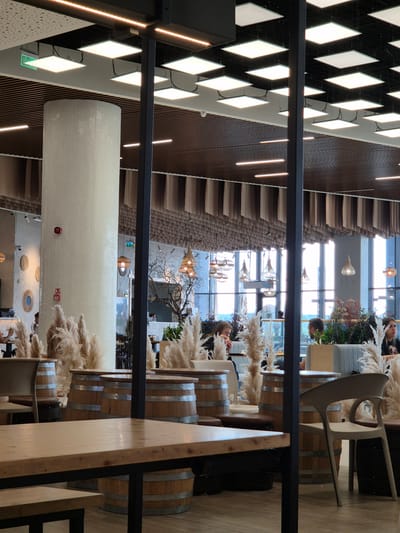 The image shows the interior of a cafe or restaurant in Craiova, Romania. The perspective is unusual, appearing to be taken from an elevated position looking down. The ceiling features a geometric black and white diamond pattern with embedded lights, interspersed with parallel wooden slats. A large, cylindrical white mosaic-tiled structure hangs from the ceiling.

The dining area features tables constructed from wooden barrels with metallic bands. Patrons are seated at some of the tables. Decorative elements include tall, feathery pampas grass arrangements placed on and around the barrels. The windows in the background provide a view of the exterior environment, suggesting an urban setting.

The image contains elements of modern design and rustic charm. The overall atmosphere seems relaxed and inviting.