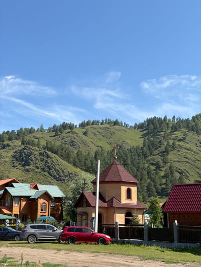 A bright, sunny day with a clear blue sky and sparse wispy clouds offers a view of a village nestled at the foot of a lush, green mountainside in Elekmonar, Russia. The mountainside is covered in a dense forest of evergreen trees, with some patches of grass and rocky outcrops visible. In the foreground, several wooden houses with distinctively colored roofs are clustered together. To the left, a multi-story wooden house with a teal roof is prominent. Next to it, a smaller wooden structure with a similar teal roof stands. In the center, a small, brick church with a reddish-brown tiled roof and a golden cross on top is visible, flanked by a wooden fence. To the right of the church, another wooden house with a dark red tiled roof is partially obscured by the church. In front of the houses and church, a dirt and grass area serves as a parking lot. Three cars are parked there: a dark gray sedan on the left, a silver SUV in the middle, and a red station wagon on the right. The scene appears peaceful and residential, with no people visible. The overall impression is one of a quiet, rural community set against a beautiful natural backdrop.