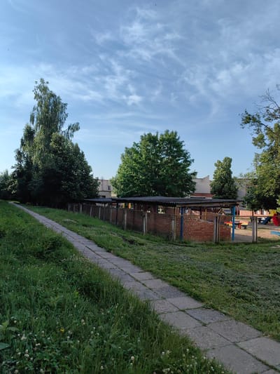 The image shows a view of a playground area on a sunny day with a clear blue sky and scattered white clouds. In the foreground, a concrete pathway runs diagonally from the bottom left corner towards the center of the image, bordered by tall green grass. Beyond the pathway, a low brick wall surrounds a structure with a corrugated roof, possibly a shelter or an unfinished building. In the background, there is a two-story building made of light-colored brick. To the right of the structure, a playground with colorful slides and benches is visible. Several trees with lush green foliage are scattered throughout the scene. The overall impression is of a recreational space within a residential or educational institution in Fanipal, Belarus. There are no people visible in the image.