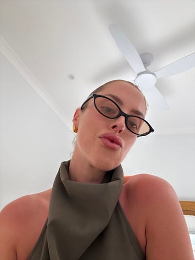 A woman looks down at the camera, wearing black-framed glasses and an olive green garment. Her lips are pursed, and a gold earring is visible on her left ear. Behind her is a white ceiling with a white ceiling fan. The viewpoint is from below, looking upwards at an angle. The background also includes a hint of wooden trim.