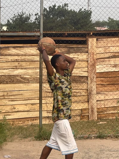 A young adult male stands in an outdoor setting in Umunekwu, Nigeria, positioned in profile facing right. He wears a multi-colored patterned short-sleeved shirt, white shorts with light blue trim, and a red wristband on his left wrist. Both hands hold a worn, brown basketball above his head in a shooting stance, arms bent at the elbows. Behind him is a chain-link fence, partially obscured by a metal pole, separating the foreground from a horizontal wooden plank structure in the mid-ground. Above the fence and wooden structure, green trees are visible against a hazy light gray sky. The ground consists of unpaved earth with scattered light green vegetation and a discarded clear plastic bottle in the lower-left.