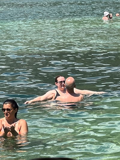 In Risan, Montenegro, three individuals are in the clear, shallow waters of the Bay of Kotor. In the foreground, a woman wearing sunglasses and an orange and white patterned swimsuit stands with her hands clasped. Further back, a couple is in the water. The man, bald and with his back to the camera, is being embraced by a woman wearing a black swimsuit and glasses. She is facing him and they appear to be in conversation. In the background, other swimmers are visible further out. The sunlit water is a beautiful shade of green, with gentle ripples reflecting the light.