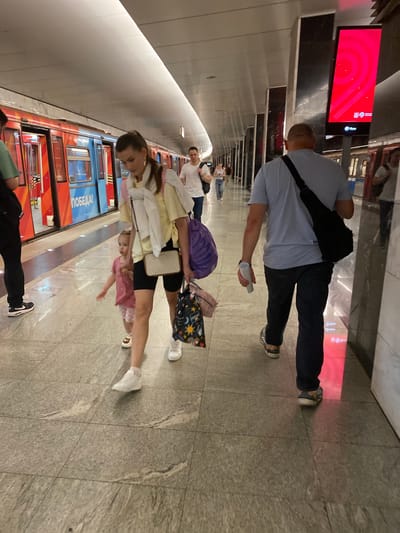 A family walks on a subway platform in Красногорск. The platform has a polished stone floor and a white ceiling with lighting. To the left, a red and blue subway train is stopped at the station, with the word "ПОБЕДА!" visible on its side. On the right, a large, vertical red digital advertisement is displayed.
In the foreground, a woman with brown hair tied in a ponytail walks to the right, carrying a purple bag over her shoulder and a tote bag with a colorful star pattern in her right hand. She wears a light yellow short-sleeved shirt, black shorts, and white sneakers. A young child, likely a girl with blonde hair, walks beside her to her left, wearing a pink t-shirt and shorts.
Further back, a man with a shaved head walks away from the camera to the right. He wears a light blue t-shirt, dark pants, and sneakers, with a black sling bag across his back. He holds a white cylindrical object in his right hand. Other individuals are visible in the background, walking on the platform. The environment suggests a public transportation hub.