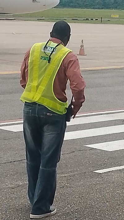 A person wearing a pink shirt, blue jeans, and a bright yellow reflective vest is standing on an airport tarmac. The vest has a black band with the text "FAA" and "OPERATIONS" visible. The person is wearing headphones and holding a dark-colored object, possibly a radio or camera, in their right hand. They are standing on a crosswalk with white stripes. In the background, part of an airplane is visible on the left, and an orange traffic cone stands on the tarmac to the right. The setting appears to be an outdoor, daytime environment at an airport, with a slightly overcast sky suggested by the lighting. The person's stance and focus suggest they are performing a duty related to airport operations.