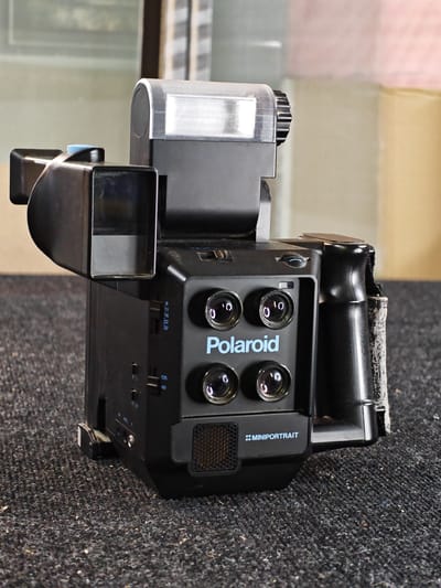 A black Polaroid Minolta camera, with a flash attachment, is displayed on a textured, dark grey surface. The camera features multiple lenses on its front panel, along with brand markings. A black handle is attached to the right side of the camera. The background is blurred, showing hints of muted colors and vertical lines, suggesting an indoor setting with soft lighting. The camera is positioned in the foreground, taking up a significant portion of the frame. The overall impression is one of a product photograph, highlighting the camera. The specific location in Ждановіцкі сельскі Савет, Belarus is not discernible from the visual information in the image.