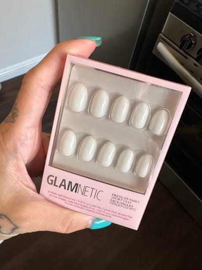 A hand holds a pink box of "Glamnetic" brand press-on nails with a clear plastic front showing ten oval-shaped, white nails. The package lists details in English and French, including the quantity of nails and the inclusion of nail glue, a file, cuticle stick, and alcohol pad. The hand holding the nails has a teal-colored fingernail and a tattoo is visible on the hand. The background includes an oven and dark-colored cabinetry. The scene appears to be indoors, possibly in a kitchen. The location is Seal Beach, United States of America.