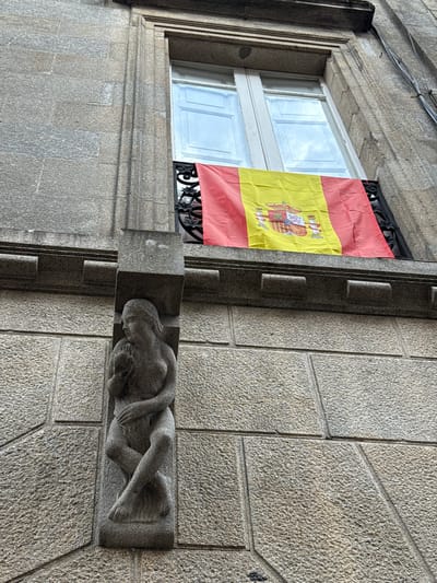 The image shows the facade of a building with a Spanish flag draped across a window. The building is constructed of granite blocks. To the left of the flag, a stone carving of a woman holding a child is visible. The carving is integrated into the building's architecture, serving as a decorative corbel. The flag is red and yellow, with the coat of arms of Spain in the center. The window above the flag appears to be a double-hung sash window with white paint. The stone work of the building is rough and textured. This scene is located on the exterior of a building in the historic center of Santiago de Compostela, Spain.